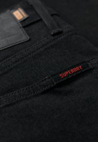 Jeans en denim noir avec une surface texturée, dotés d'une patch en cuir noir et d'une étiquette rouge portant la mention "SUPERDRY."