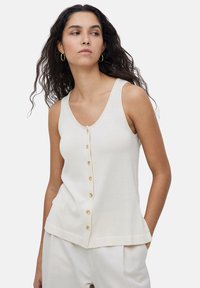 Top de punto de color crema claro con cuello en V, que presenta seis botones de madera en el frente y un dobladillo recto. Diseño minimalista, estilo sin mangas.
