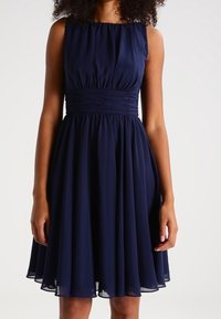 Robe sans manches bleu marine avec un corsage ajusté et une taille froncée, présentant une jupe fluide en tissu léger et texturé.