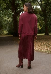 Maroon sweatshirt, plisserad maroon maxikjol och bruna klackstövlar, mot en parkbakgrund med gröna träd och fallna löv.