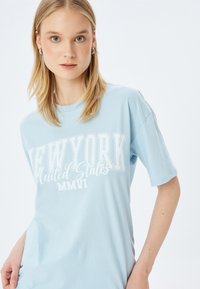 Lichtblauwe katoenen T-shirt met korte mouwen, met in het vet gedrukte witte tekst "NEW YORK Verenigde Staten MMVI" in verschillende lettertypen.