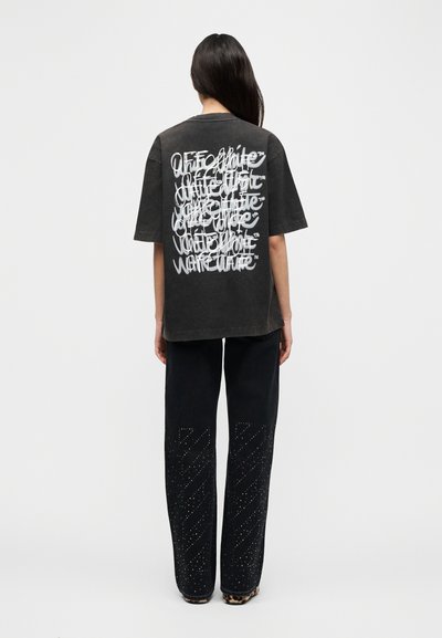 Μαύρο φαρδύ t-shirt με λευκό κείμενο σε στυλ γκράφιτι στην πλάτη, συνδυασμένο με μαύρο παντελόνι που έχει διαγώνια σχέδια με πριτσίνια.