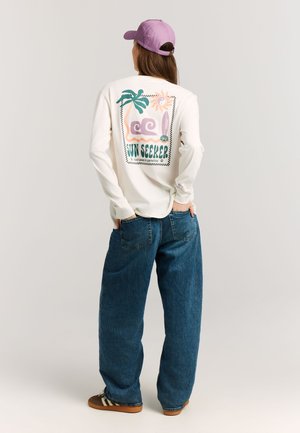 Persoon die een wit shirt met lange mouwen draagt met een kleurrijk "Sun Seeker" strandontwerp, blauwe wijde jeans, bruine sneakers en een paarse pet, met de rug naar voren gekeerd.