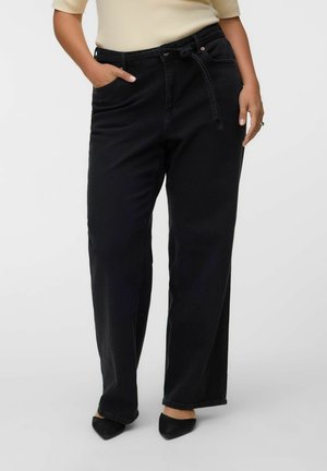 Kvinde iført sorte jeans med høj talje og vide ben med taljebånd, beige top og sorte spidse sko, stående med den ene hånd i lommen.
