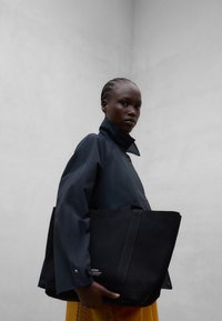 Personne avec des cheveux tressés portant une veste foncée à col montant et tenant un grand sac fourre-tout noir sur un fond uni gris clair.