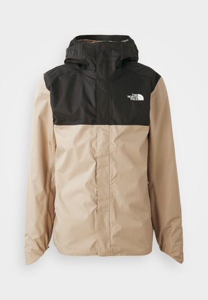 Casaco corta-vento em bege e preto com capuz, apresentando um fecho zip frontal e bolsos laterais. Logótipo na área do peito esquerdo. Textura suave.