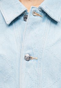 Κοντινό πλάνο ανοιχτού μπλε μπουφάν από τζιν με ασημένια κουμπιά snap Calvin Klein και μπεζ κουμπωματικές τρύπες, που δείχνει τον γιακά και το πάνω μέρος του στήθους.