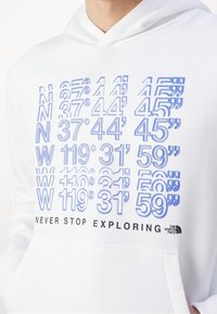 Sudadera blanca con texto azul que muestra coordenadas y "NUNCA DEJES DE EXPLORAR" en negro, con el logo de The North Face en la esquina inferior derecha.