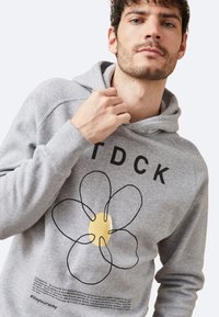 Sudadera con capucha gris que presenta un diseño de flor negra con un centro amarillo, acentuado por letras en negrita "TDCK" arriba y texto debajo. Textura suave.