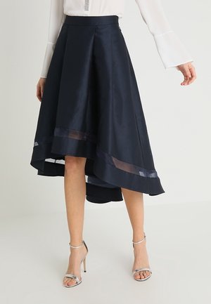 Femme portant une jupe asymétrique bleu marine avec des rayures horizontales transparentes près de l'ourlet, associée à des sandales à talons hauts argentées à brides.