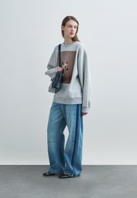 Moteris dėvi pilką džemperį su šuns atspaudu, plačias mėlynas džinsines kelnes su šoninėmis juostelėmis, juodus batus ir laiko juodą peties krepšį.