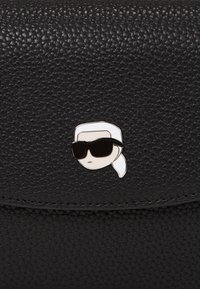 Geantă din piele texturată neagră, decorată cu un pin din email de culoare argintie reprezentând un chip cu ochelari de soare și un batic alb pe față.