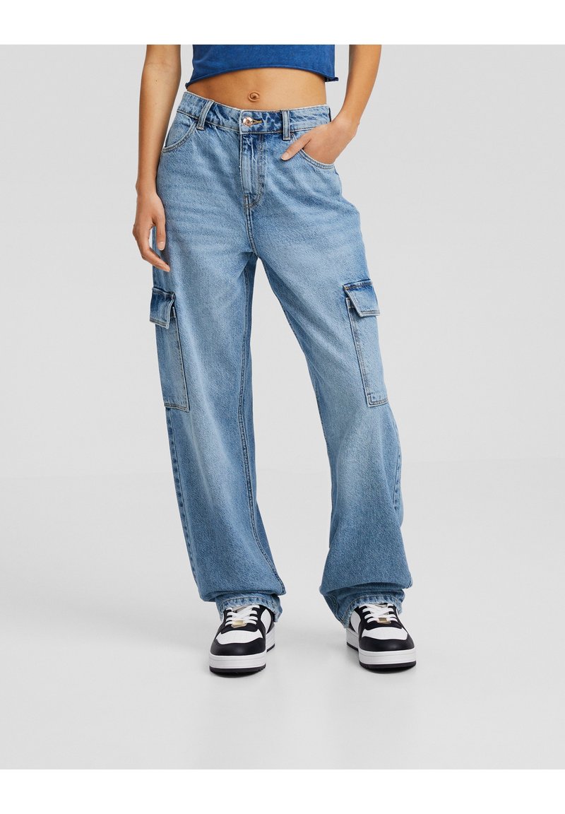 High-waisted, light blue denim cargo jeans with wide legs, featuring side pockets and a zipper fly. Worn with black and white sneakers.