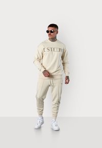 Sweatshirt beige et jogging avec logo "ESTEEM", mélange de coton, poignets côtelés, tons assortis, associés à des sneakers blanches et des lunettes de soleil noires.