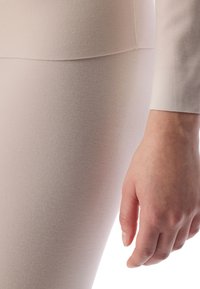 Hellbeige, strukturiertem Stoff, der ein enganliegendes Kleidungsstück bildet, mit einer Nahaufnahme einer Hand, die dagegen ruht. Glatte, minimale Nähte sichtbar.