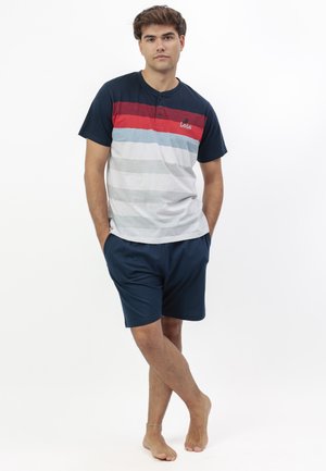 Joven de pie descalzo con las manos en los bolsillos, vistiendo pantalones cortos azul marino y una camisa de manga corta con rayas multicolores.