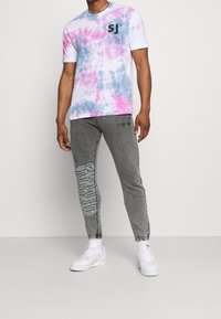 Tričko s krátkým rukávem ve stylu tie-dye v bílé, růžové a modré barvě s černým logem. Šedé joggers kalhoty s bílým textovým vzorem, ribbenými manžetami a bílými teniskami.