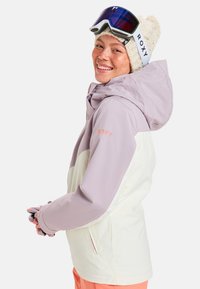 Ski jacket with a lavender and white design, featuring a hood, zip pockets, and Roxy logo. Model wears a knitted hat and ski goggles.