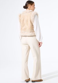 Vrouw met de rug naar de kijker draagt een beige nepbont vest over een witte blouse met lange mouwen, crèmekleurige wijde broek en bruine schoenen, op een witte achtergrond.