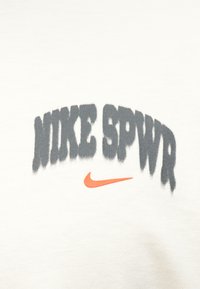 Balta t-kreklā ir redzams teksts "MIKE SPVR" tumšajā pelēkā krāsā un zem tā oranžs Nike swoosh logo, kas drukāts treknā un stilizētā fontā.