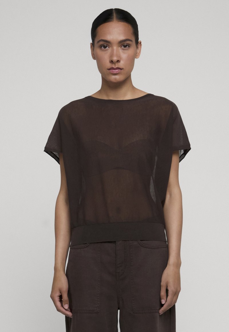 Femme aux cheveux foncés avec une raie au milieu porte un haut noir transparent à manches courtes sur un soutien-gorge noir et un pantalon foncé, debout devant un fond uni.