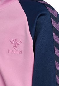 Hummel-Sportoberteil mit pinkem Stoff, marineblauen Ärmeln und strukturierten lila Chevron-Akzenten. Auf der Brust ein gesticktes Logo.