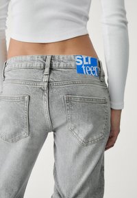 Sive traperice s visokim strukom, s plavom oznakom koja kaže "SLI teen", dva stražnja džepa i izbljedjela tekstura.