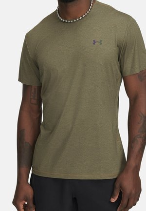 Mężczyzna ubrany w oliwkową, sportową koszulkę z krótkim rękawem z małym logo Under Armour na lewej piersi i czarne spodenki.