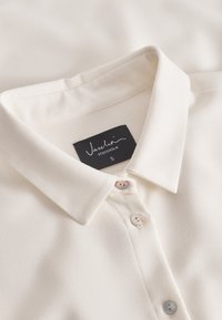 Bluză albă cu guler, fabricată dintr-un material neted. Are o închidere cu nasturi perle și o etichetă neagră inscripționată "Jascha, Stockholm, S."