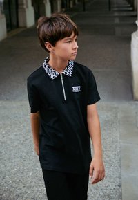 Polo nera con colletto bianco decorato e zip frontale, con un piccolo logo sul petto. Indossata con pantaloni neri.