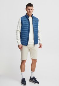 Junger Mann trägt eine blaue gesteppte Weste, weißen Pullover, beigen Shorts, weiße Socken und dunkelblaue Turnschuhe, steht vor einem einfarbigen Hintergrund.