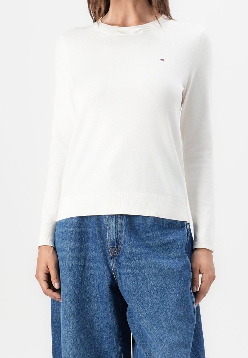 Femme portant un pull blanc à manches longues avec un petit logo sur la poitrine et un jean bleu ample, debout devant un fond blanc.