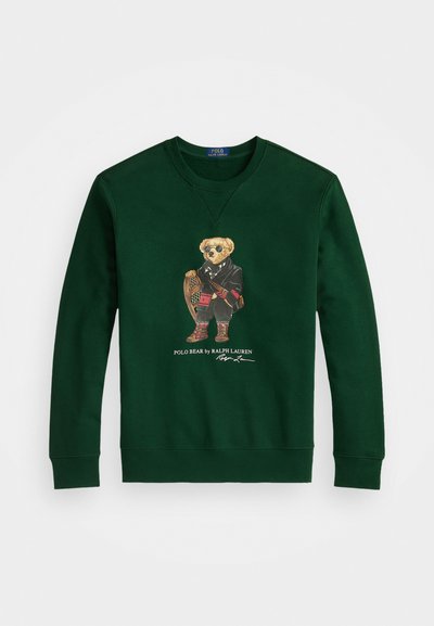 Zaļš kokvilnas džemperis ar lāci žaketē un saulesbrillēs, kas tur sniega kurpes, zem kura ir teksts "Polo Bear by Ralph Lauren".