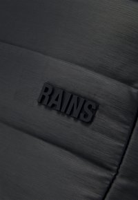 Surface texturée noire avec des crêtes horizontales présentant un logo "RAINS" en relief avec des lettres noires en gras.
