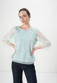 Blusa trasparente azzurro chiaro con dettagli di frange di perline intricate, scollatura rotonda e maniche a tre quarti, abbinata a jeans scuri.