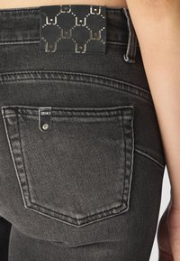 Svarte denimjeans med baklomme. Et merke med geometrisk mønster og gullacenter er synlig på livet. Strukturert stofffinish.
