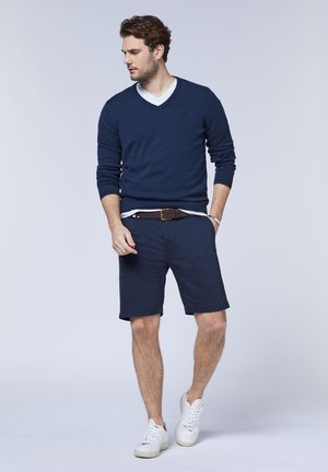 Marineblaue V-Ausschnitt-Pullover über einem weißen Hemd, kombiniert mit marineblauen Shorts und weißen Sneakers. Der Gürtel ist aus braunem Leder und der Stoff wirkt glatt.