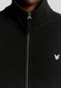 Zwarte zip-up jack met hoge kraag en zilveren hardware. Heeft een klein wit adelaarlogo op de linkerborst.