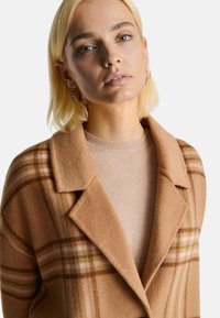 Kamelowy wełniany płaszcz w kratę w odcieniach beżu i zieleni. Posiada wykładany kołnierz i przytulny beżowy sweter pod spodem.