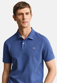 Muž nosící modré polo tričko krátký rukáv s malým vyšívaným logem na levé hrudi, hledící mírně stranou.