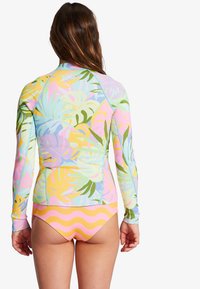 Camiseta de manga larga con protección solar en colores pastel y patrones de hojas tropicales. Combinada con un bikini de tiro bajo con rayas amarillas y rosas.