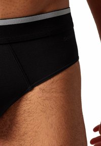 Brief in cotone nero con elastico argentato in vita, caratterizzato da una texture liscia e un design aderente. La vista laterale mostra i peli corporei e il tono della pelle.