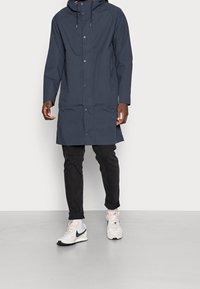 Man i en marinblå huva-regnjacka, svarta uppnosade byxor, vita sneakers med svart Nike-logotyp, stående på ett ljusgrått golv.