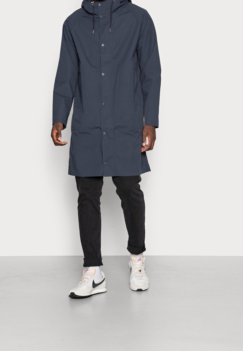 Man i en marinblå huva-regnjacka, svarta uppnosade byxor, vita sneakers med svart Nike-logotyp, stående på ett ljusgrått golv.