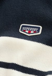 Gestrickter Stoff mit navyblauen und weißen horizontalen Streifen, der ein aufgenähtes Patch mit dem Text "SUPERDRY" sowie roten und weißen Streifen aufweist.