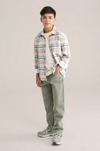 Rutig flanellskjorta i beige, blått och brunt över en krämfärgad t-shirt. Olivgröna byxor och ljusgrå sneakers med svarta accenter.