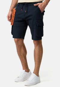 Marine cargo shorts med snor, flere lommer og orange mærkedetalje. Båret med hvide sneakers. 100% bomuldsstof, afslappet pasform, knælængde.
