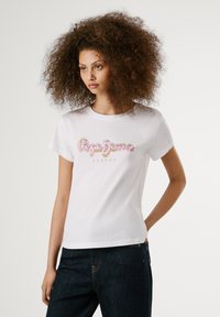 T-shirt en coton blanc arborant un logo "Pepe Jeans London" en rose et or sur la poitrine. Manches courtes et encolure ronde.
