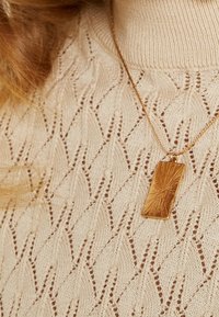 Beżowy sweter dzianinowy z teksturowym wzorem oraz złotym naszyjnikiem w kształcie prostokąta, zdobionym wzorem promieni słonecznych.