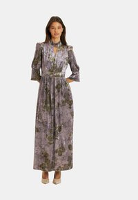 Largo vestido de terciopelo morado con un patrón floral, con mangas abullonadas, un escote alto y una cinturilla. Combinado con tacones de color claro.